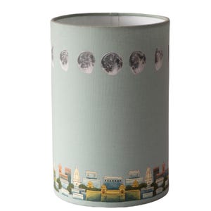 May include: A light blue lampshade with a cityscape and moon phases design. The cityscape is painted in shades of yellow, orange, and blue. The moon phases are silver and are arranged in a row at the top of the lampshade.