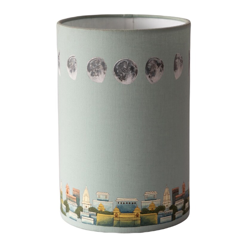 May include: A light blue lampshade with a cityscape and moon phases design. The cityscape is painted in shades of yellow, orange, and blue. The moon phases are silver and are arranged in a row at the top of the lampshade.