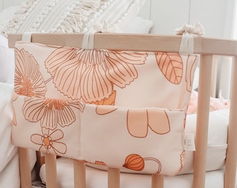 Peut inclure: Un lit d'appoint en bois pour bébé avec un matelas blanc et un tissu à motifs floraux suspendu sur le côté. Le lit d'appoint est fixé à un cadre de lit blanc.