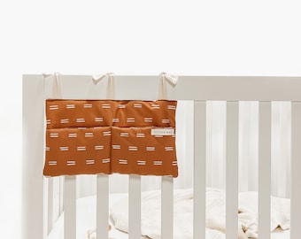 Peut inclure: Un lit bébé blanc avec un organisateur suspendu à motifs marrons et blancs avec le texte "Little Wag" dessus. L'organisateur a trois poches.