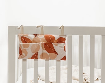 Peut inclure: Un lit bébé blanc avec un drap crème et un organisateur suspendu avec trois poches. L'organisateur a un motif floral dans les tons orange, pêche et blanc. Le texte "Little & Mai" est imprimé sur l'organisateur.