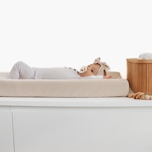 May include: A beige changing pad with a baby lying on it. The baby is wearing a white onesie and a brown headband. The changing pad is on a white changing table. There is a bamboo basket with a white towel in it and a wooden toy on the table.