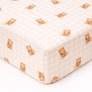 May include: A fitted crib sheet featuring a light beige and white gingham check pattern. The fabric is decorated with a repeating print of small, brown teddy bears in a seated position, evenly spaced across the entire surface of the textile.