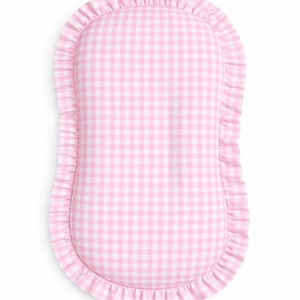 May include: An oval-shaped cushion featuring a light pink and white gingham check pattern. The perimeter of the cushion is finished with a gathered fabric ruffle in the same pink and white checkered print, creating a soft, textured border.
