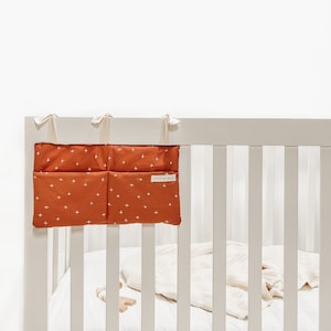 May include: A white crib with a white and orange crib organizer with the text "Little Man" on it. The organizer has three pockets and is hanging on the side of the crib.