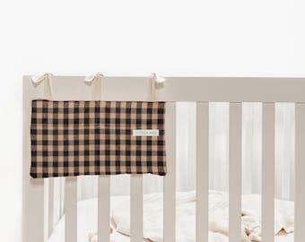 Peut inclure: Une protection de barre de lit à carreaux noirs et beiges avec le texte "LITTLE MAE" sur une étiquette blanche. La protection est fixée à un lit bébé blanc avec des liens crème.