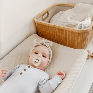 May include: A baby wearing a light blue onesie and a matching bow headband lies on a beige changing pad. A pacifier is in the baby's mouth. A woven basket with a blanket is in the background.