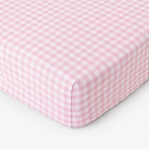 May include: A fitted sheet with a pink and white gingham pattern. The fabric is folded over at the corner, showing the texture and pattern of the material. The sheet is made of a soft, breathable fabric.