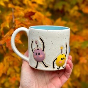 May include: Speckled white ceramic mug with cartoon characters in purple and yellow with black outlines. The inside of the mug is light blue. The mug has a curved handle. The mug is suitable for hot drinks.
