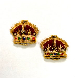 Pode incluir: Duas coroas bordadas a ouro com centros de veludo vermelho e detalhes de joias. As coroas estão sobre um fundo branco.
