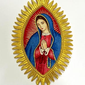 May include: An embroidered patch featuring a figure with downcast eyes and hands pressed together in prayer. The figure wears a blue mantle over a red robe, set against a red background within a golden, sunburst-shaped oval frame with rays.
