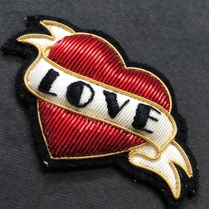 May include: A red heart with gold trim and a white ribbon that says "LOVE" in black embroidery. The heart is a traditional tattoo design.