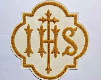 May include: An off-white fabric patch featuring gold embroidered lettering of the letters IHS with a cross extending from the top of the H. The design is enclosed within a gold embroidered quatrefoil border on a cream-colored background.