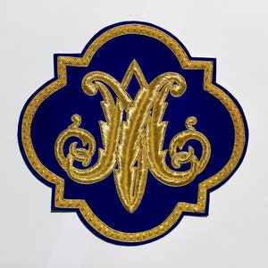 May include: A royal blue quatrefoil-shaped patch featuring an intricate gold metallic thread embroidery of the letter M in a stylized script font. The border of the patch is also outlined with a detailed gold metallic thread trim.