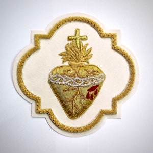 Puede incluir: Un parche decorativo con un diseño bordado del Sagrado Corazón en hilo dorado sobre una base de tela blanca. El corazón está rodeado por una corona de espinas plateada, rematado con una cruz y llamas doradas, y acentuado con tres gotas rojas.