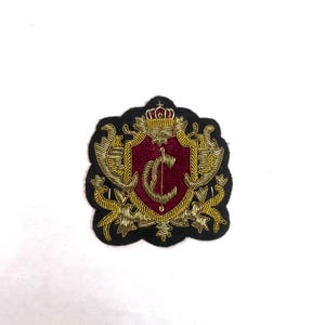 Puede incluir: Parche bordado con diseño de escudo. Presenta un escudo burdeos con la letra "C" dorada, rodeado de adornos dorados y un borde negro. Una corona dorada corona el escudo.