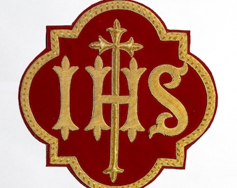 May include: A red, quatrefoil-shaped patch featuring gold-colored metallic embroidery. The design displays the letters IHS in a stylized font, with the letter H integrated into a cross. The entire emblem is outlined with a gold-colored border.