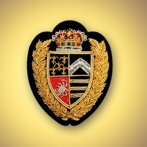 May include: A shield-shaped embroidered patch featuring a gold crown with colourful accents at the top. The central shield is divided into four quadrants displaying gold fleur-de-lis, a silver chevron, a silver crab, and horizontal silver bars.