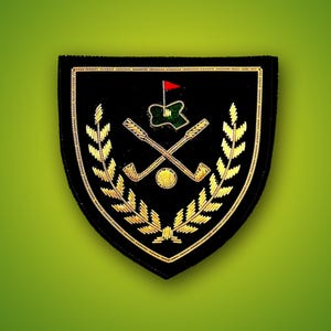 Può includere: Una toppa nera a forma di scudo con foglie di alloro ricamate in oro che incorniciano due mazze da golf incrociate e una pallina da golf. Sopra le mazze c'è un piccolo green verde con una bandierina rossa. Il tutto è bordato in filo d'oro.