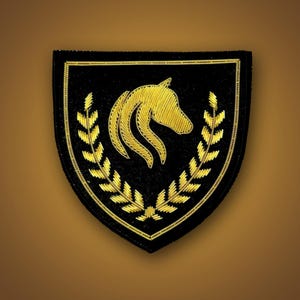 May include: A shield-shaped patch featuring black fabric with gold embroidered details. The design includes a central horse head profile facing right, flanked by two vertical laurel leaf branches, all enclosed within a thin gold border.