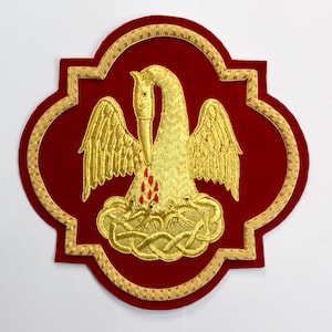 May include: A deep red velvet patch featuring an embroidered gold pelican in its piety motif. The design shows a golden pelican feeding its young, surrounded by a decorative gold braided border in a quatrefoil shape with intricate gold stitching.