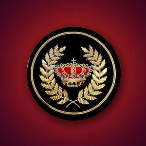 May include: A circular black patch featuring gold metallic thread embroidery in the shape of a laurel wreath surrounding a central crown. The crown is detailed with red thread and small green and blue accents, all enclosed by a gold circular border.