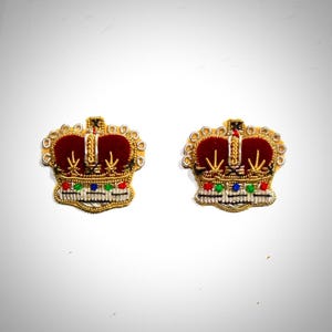 Puede incluir: Dos parches de corona bordados en oro con centros de terciopelo rojo y detalles plateados. Las coronas tienen un diseño tradicional con una parte superior puntiaguda y una base circular.