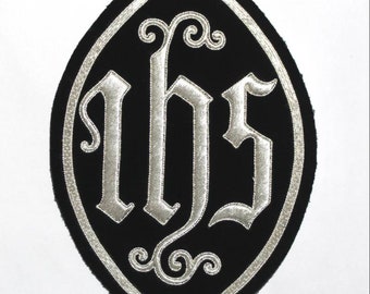 May include: An oval-shaped black fabric patch featuring the silver embroidered letters IHS in a gothic script. The letters are centered within the patch, which is bordered by a silver embroidered line that follows the pointed oval contour.