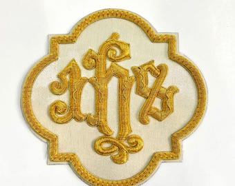 May include: A quatrefoil-shaped embroidered patch featuring a cream-colored fabric base with a gold-colored border. The center displays the gold-colored embroidered monogram IHS in a stylized, ornate script with decorative flourishes.