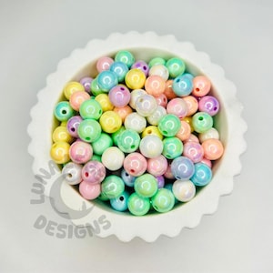 May include: A white, scalloped bowl filled with colourful, iridescent beads. The beads are a mix of pastel colours including pink, green, yellow, and blue. The beads are round and have a glossy finish. The bowl is on a white surface.