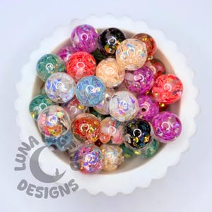 Könnte beinhalten: Eine Sammlung bunter, runder Perlen mit schillerndem Glitzer und floralen Akzenten. Die Perlen sind in verschiedenen Farben erhältlich, darunter Rosa, Lila, Grün und Orange, und werden in einer weißen Schale mit gewelltem Rand präsentiert. Das Luna Designs-Logo ist sichtbar.
