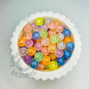 May include: A white, scalloped-edge bowl filled with colorful, translucent beads. The beads are various colors, including orange, pink, blue, green, and yellow. The beads have a glittery, iridescent appearance. The bowl is on a white surface.