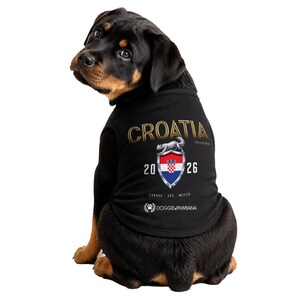 May include: A Rottweiler dog wearing a black shirt with "CROATIA" in gold lettering. The shirt features a shield with the Croatian flag and the year "2026". The shirt also has the text "DOGGIEPAWBANA" and the locations "CANADA USA MEXICO".