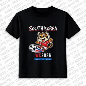 May include: Black t-shirt featuring a cartoon tiger kicking a soccer ball. The shirt has the text "SOUTH KOREA" above the tiger and "WC2026" below, with "CANADA USA MEXICO" underneath.