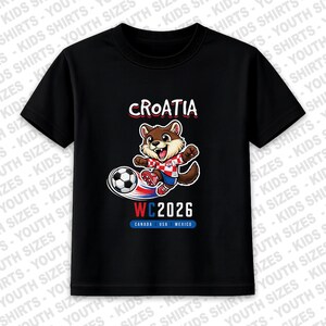 May include: Black youth t-shirt with a cartoon otter kicking a football. The shirt says "CROATIA" in red and white, with "WC2026" and the flags of Canada, USA, and Mexico. A playful design for young football enthusiasts.