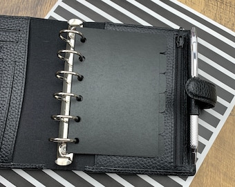 May include: Black leather planner with silver binder rings, a zippered closure, and a pen holder. The planner is open, revealing black divider pages. A silver pen is in the pen holder. The planner rests on a black and white striped paper.