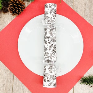 May include: A white plate with a black and white patterned Christmas cracker in the center, set on a red square. The cracker has a botanical design. Pine needles and pine cones are in the background.