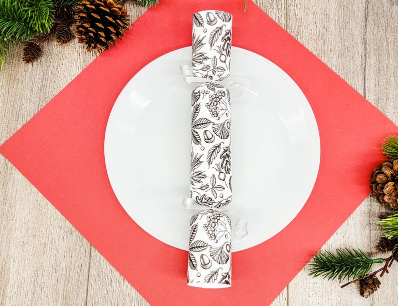 May include: A white plate with a black and white patterned Christmas cracker in the center, set on a red square. The cracker has a botanical design. Pine needles and pine cones are in the background.