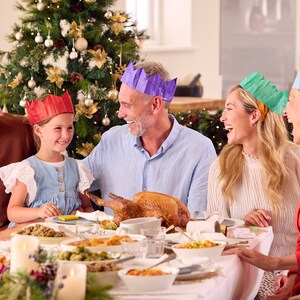 May include: A family gathers around a table set for a holiday meal. The table is laden with food, and the family members are wearing festive paper crowns. A decorated Christmas tree is in the background, adding to the celebratory atmosphere.