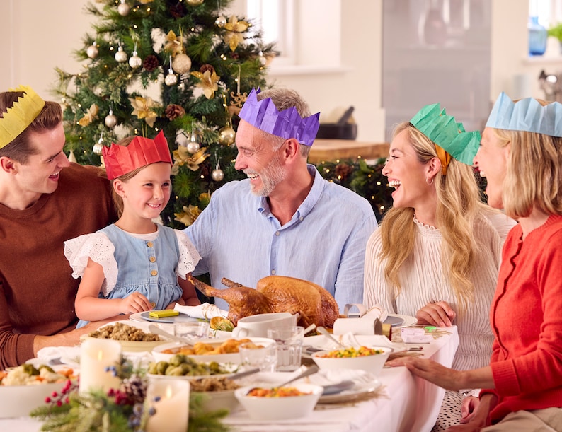 May include: A family gathers around a table set for a holiday meal. The table is laden with food, and the family members are wearing festive paper crowns. A decorated Christmas tree is in the background, adding to the celebratory atmosphere.