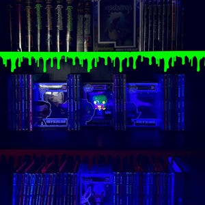 May include: A shelf featuring Goosebumps books and Funko Pop figures, lit with blue light. The spines of the books and the figures are visible. A green slime design decorates the top shelf. The image includes the text "Goosebumps".