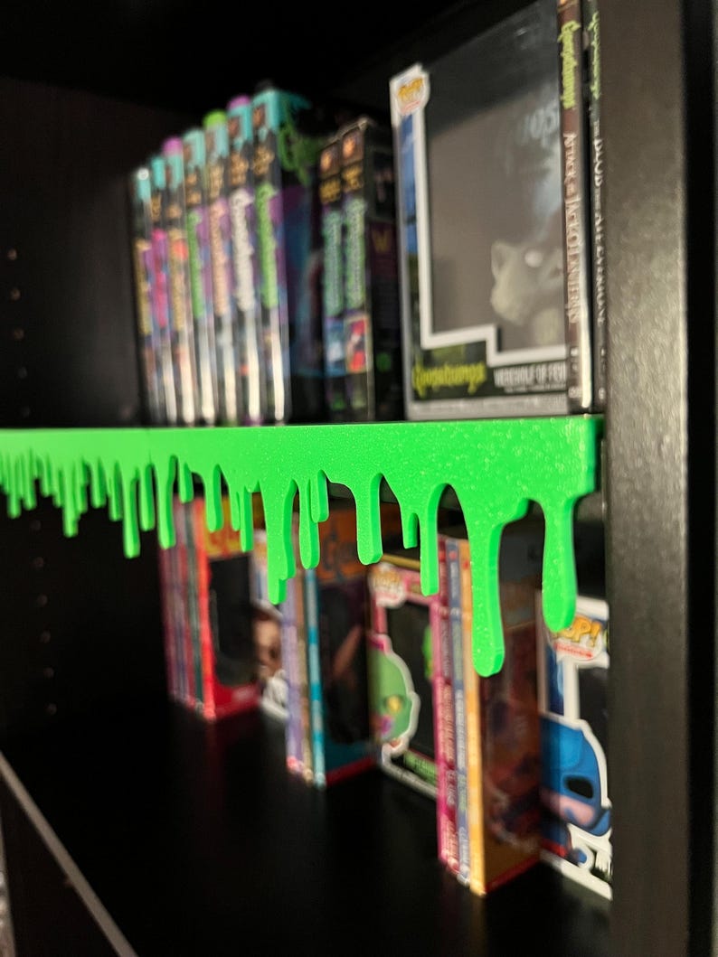 May include: A bright green shelf with a dripping slime design. The shelf is mounted on a black bookshelf, displaying Goosebumps books and Funko Pop figures. The shelf adds a unique, spooky aesthetic to the display.