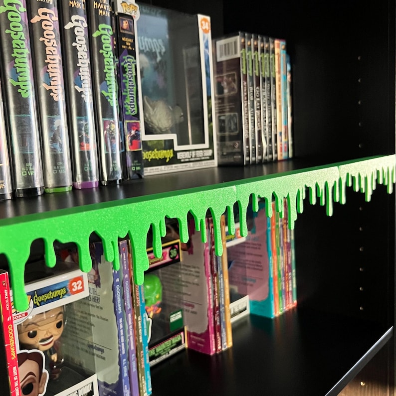 May include: A black bookshelf displaying Goosebumps books, DVDs, and collectibles. A vibrant green, dripping slime shelf border provides a decorative element. The shelf is filled with items from the Goosebumps series.