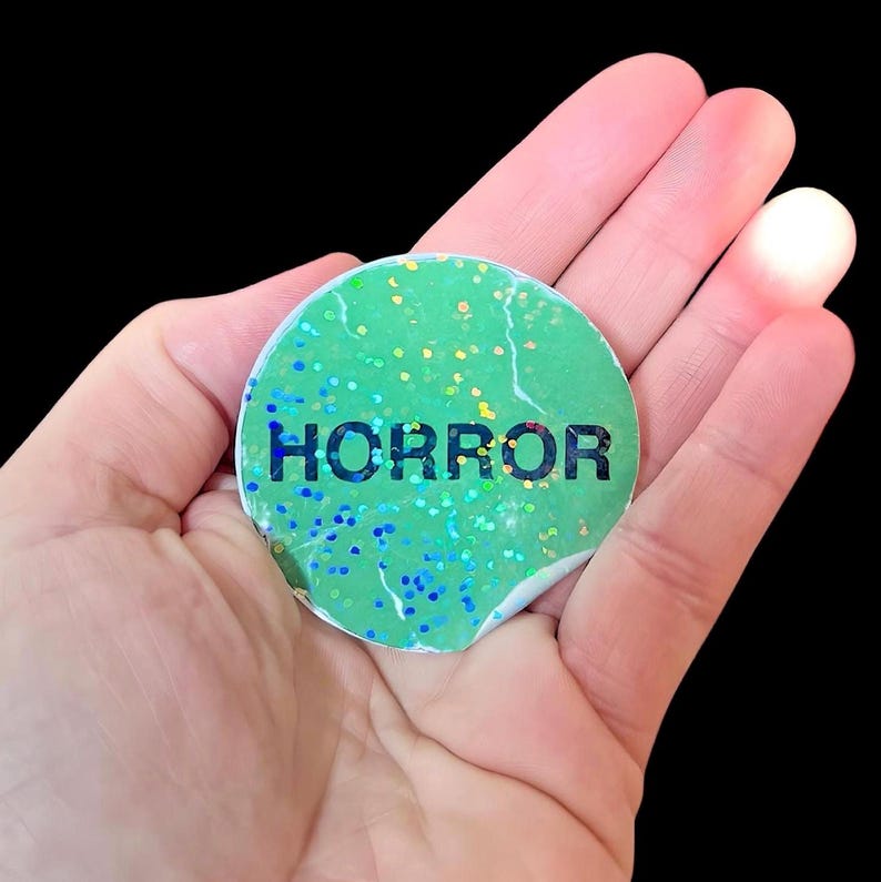 May include: A green round sticker with iridescent glitter and the word "HORROR" printed in black.