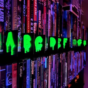 May include: A shelf of DVDs and VHS tapes is illuminated by glowing green alphabet letters. The letters A through G are displayed in a dripping font. The background features various movie titles, creating a spooky, horror-themed display.