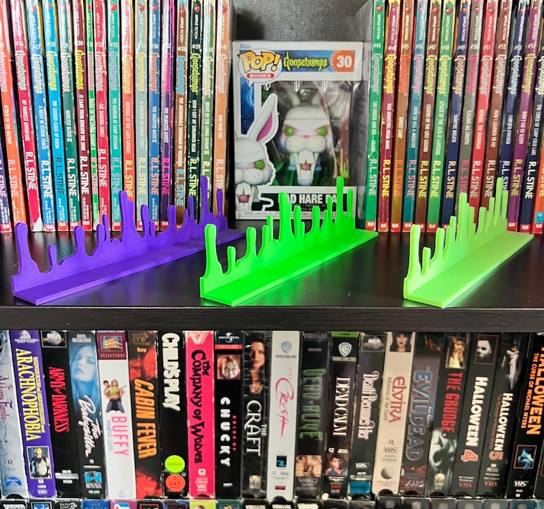 May include: Three 3D-printed bookends in the image. One is purple, and two are green. The bookends have a dripping slime design. The bookends are on a shelf with Goosebumps books and VHS tapes.