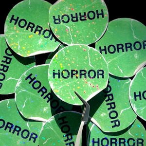 May include: A close-up of several green round stickers with the word "HORROR" printed in black letters. The stickers have a glittery, speckled texture.