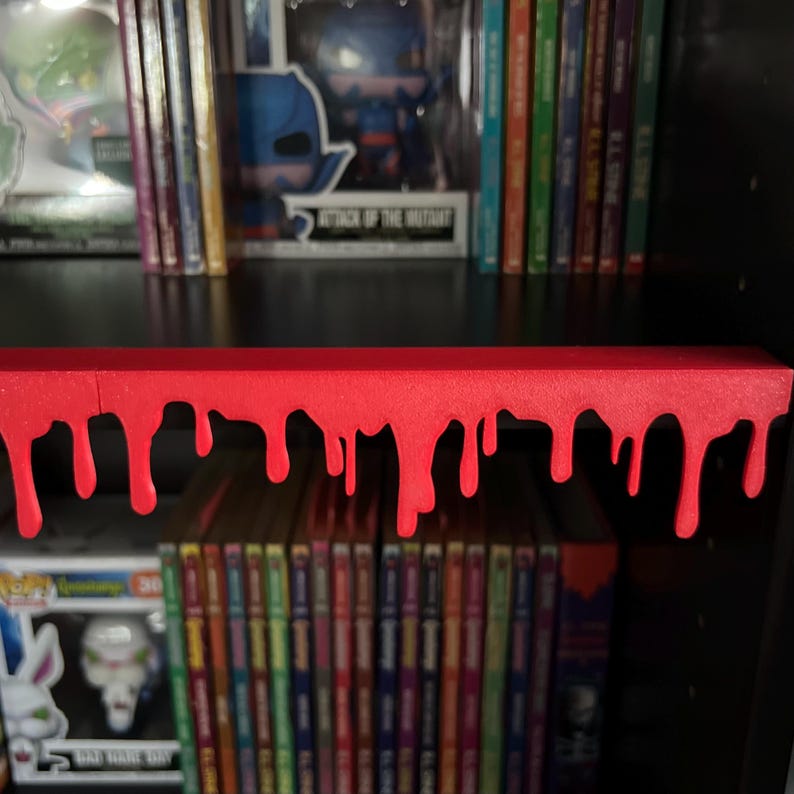 May include: A red shelf with a dripping blood design. The shelf is positioned in front of a collection of books and figurines. The shelf is a decorative element, adding a spooky or horror theme to the display.