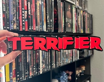 Puede incluir: Un letrero 3D impreso en rojo y negro con la palabra "TERRIFIER" en letras rojas y negritas. El letrero se sostiene frente a una estantería llena de DVD.