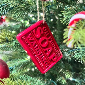 May include: A red rectangular Christmas ornament with the words "OH FUDGE SOAP WARNING MAY CAUSE DUMBNESS" in raised letters. The ornament hangs from a twine string on a Christmas tree with other ornaments.
